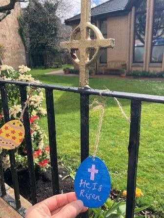 A blue paper Easter egg on the railings that says "He is Risen", with a stone cross behind.